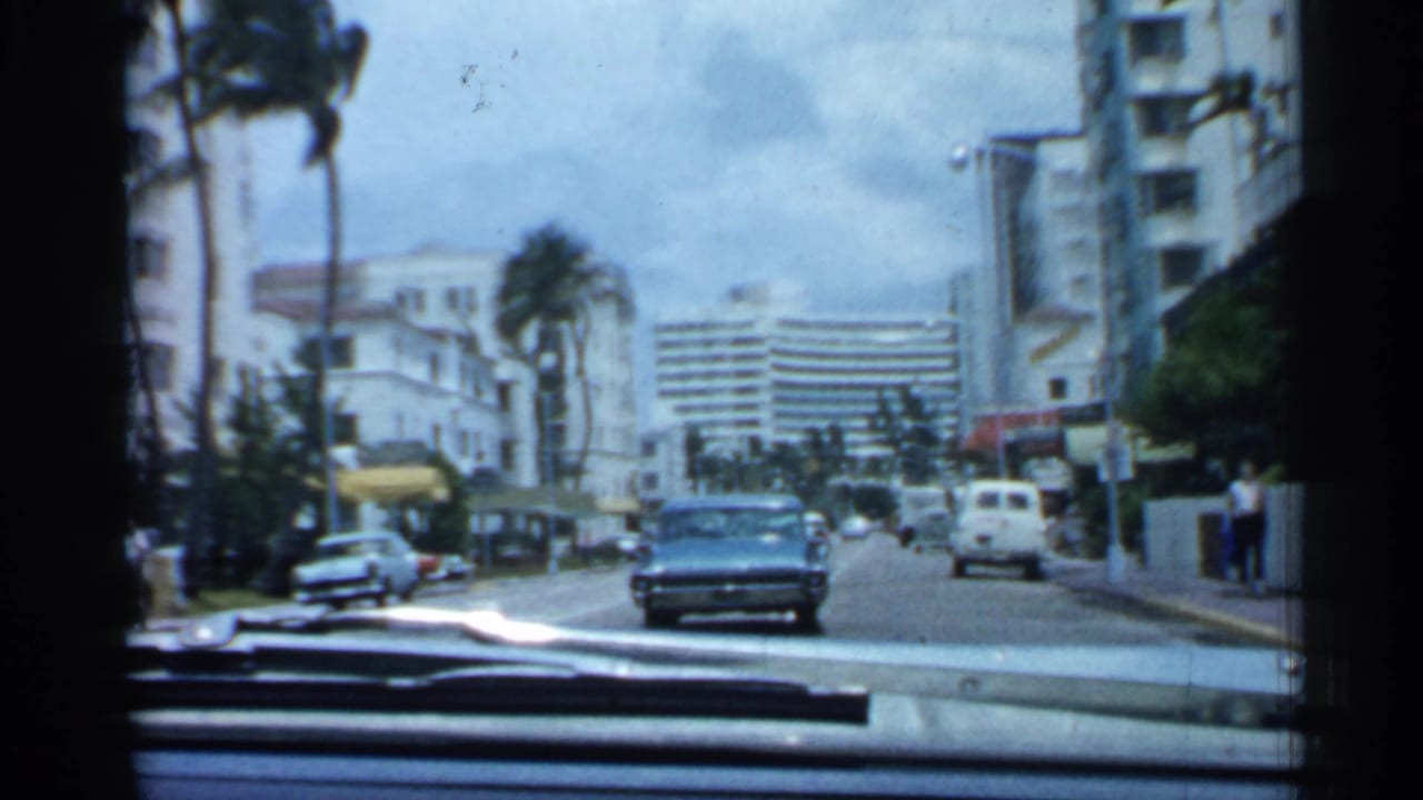 Windshield view down a city street with cars, palms, white buildings, and a blue sedan ahead.
