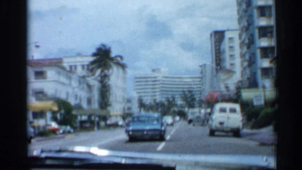 Moving car view of a boulevard with a blue sedan, white van, palms, and tall buildings.
