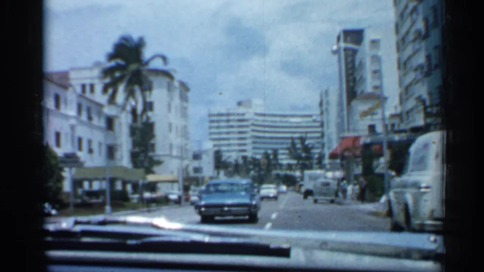 1958 Florida street-driving frame showing a blue sedan ahead, storefront awnings, palms, parked cars, pedestrians, and high-rise buildings.