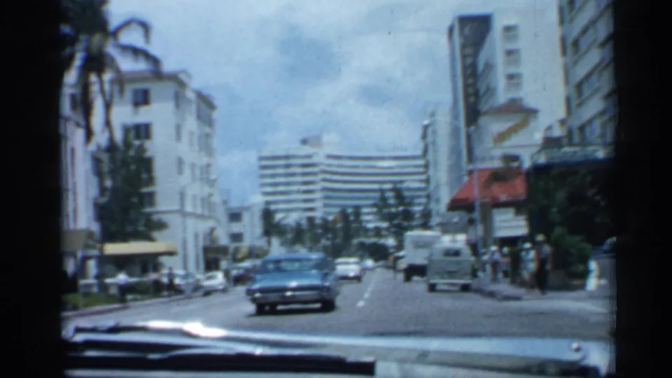 1958 Florida boulevard view from inside a moving car, with a blue sedan, pedestrians on the sidewalk, palm trees, awnings, and white city buildings.