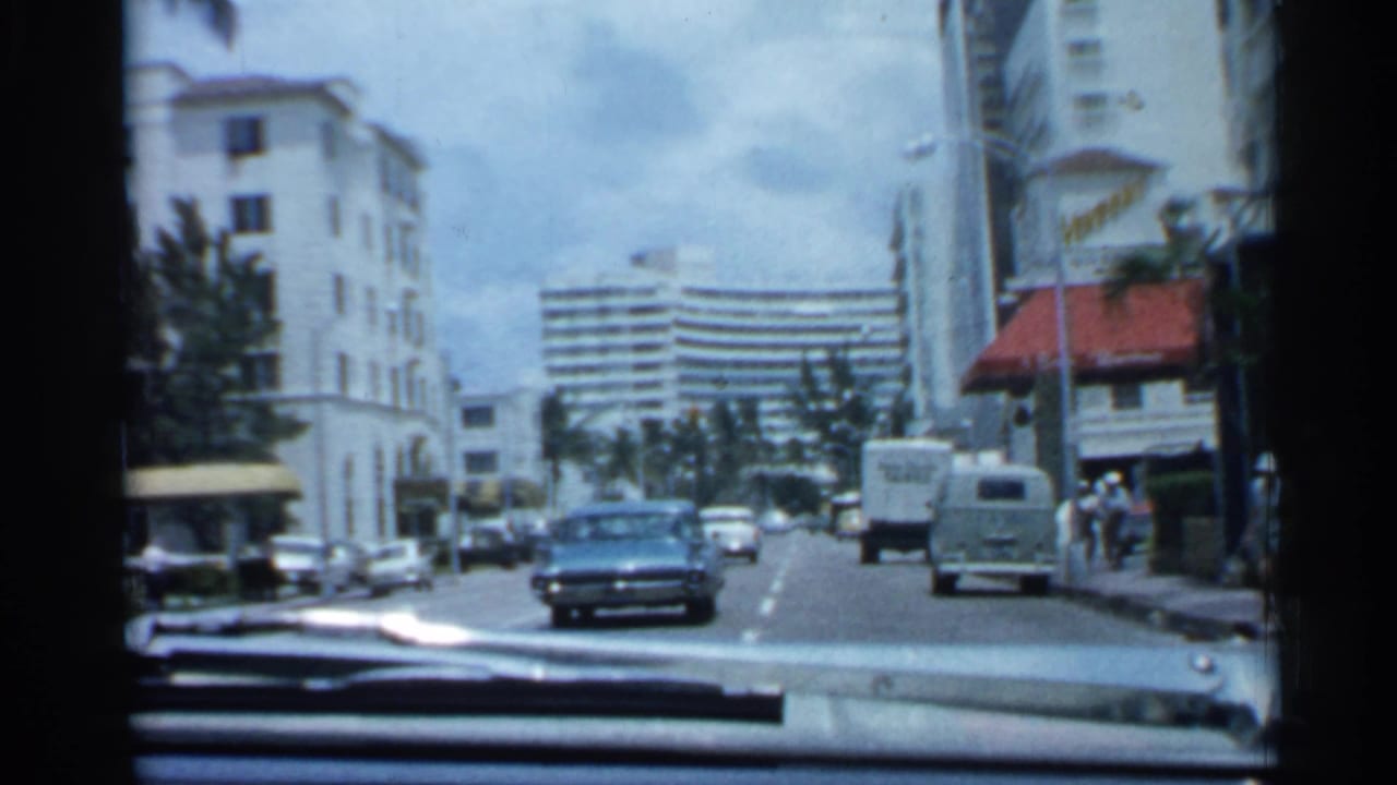 1958 Florida home-movie view from a car on a city street, with a blue sedan ahead, mid-century buildings, palms, and traffic.