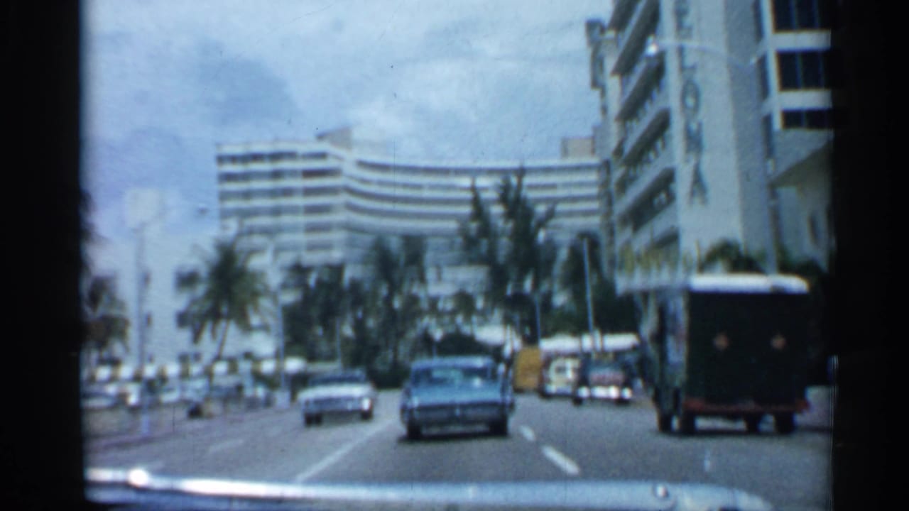 1958 Florida street-driving frame showing a blue sedan, other cars, palm trees, and a large curved hotel or apartment building ahead.