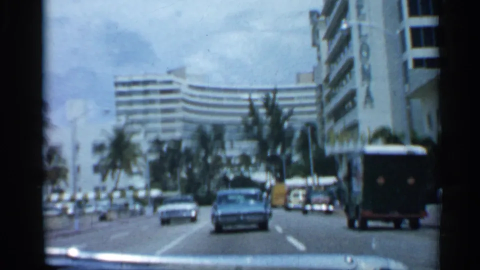 1958 Florida street-driving frame showing a blue sedan, other cars, palm trees, and a large curved hotel or apartment building ahead.