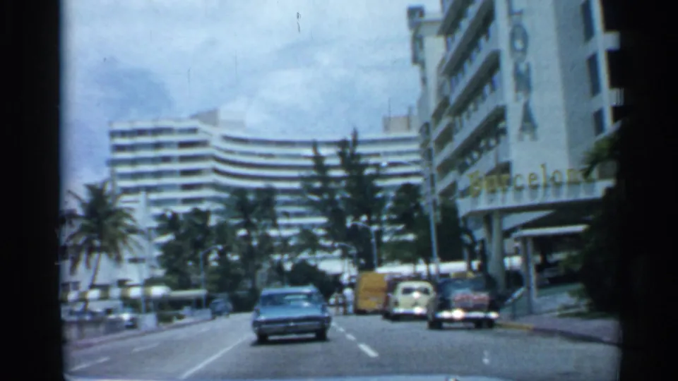 1958 Florida urban road scene with a blue sedan ahead, palms, parked vehicles, pedestrians, and visible building signage at right.