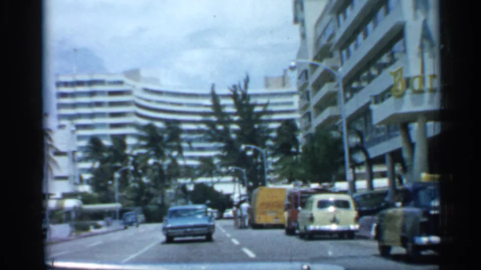 1958 Florida city-street frame with a blue sedan, curbside cars, pedestrians, palms, and modern buildings along the road.
