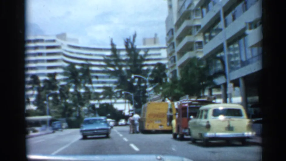 1958 Florida home-movie view down a city street with palms, multi-story buildings, parked cars, a yellow truck, and cars ahead.