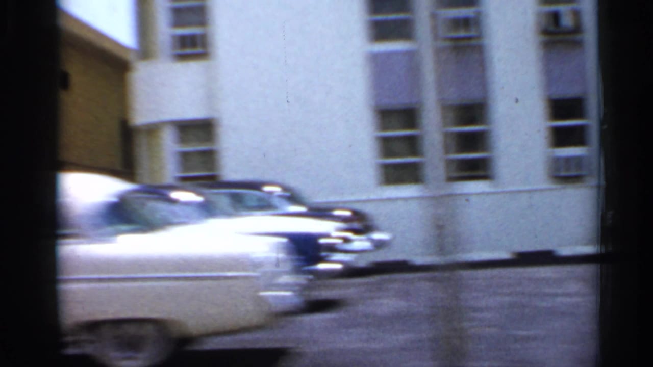 Blurred white car passing in front of a light-colored building with windows and wall air conditioners.