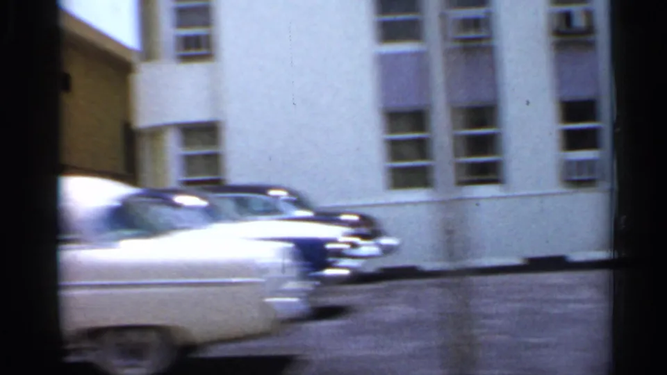 A blurred mid-century car passes a light-colored urban building during the 1958 Florida street-driving footage.