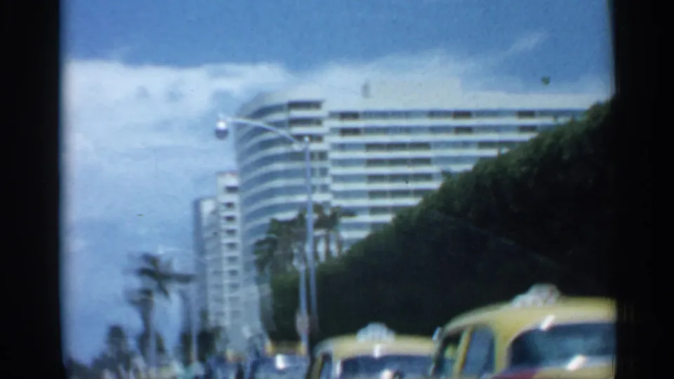 1958 Florida street view with a large curved mid-century building, palms, streetlights, hedges, and yellow vehicles at the curb or in traffic.