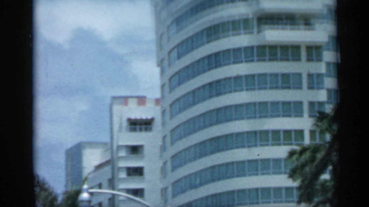 1958 Florida 8mm frame showing a rounded high-rise facade, window bands, palm trees, and a partly cloudy sky.