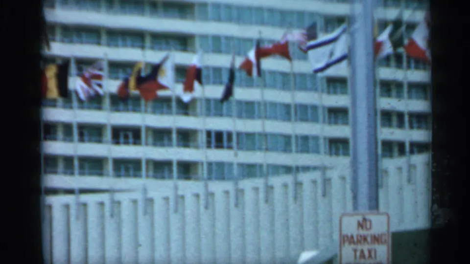 1958 Florida 8mm frame showing international flags along a high-rise facade and a No Parking Taxi sign near the lower right.