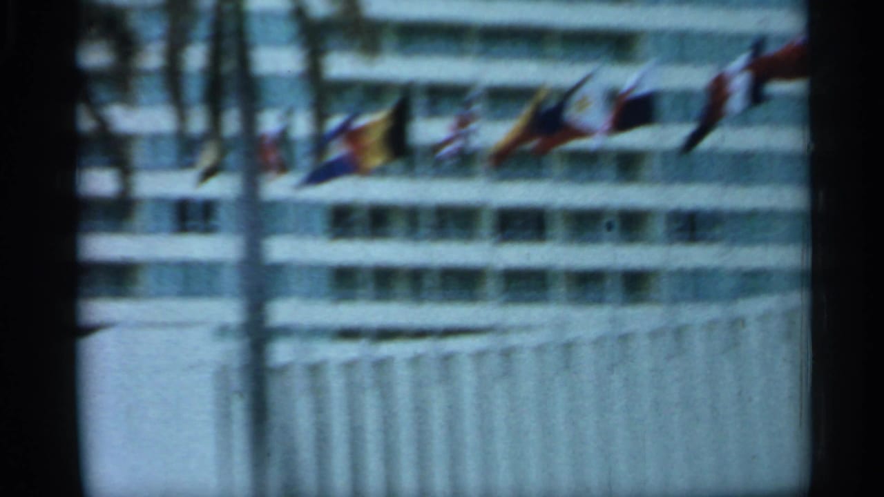 1958 Florida home-movie frame showing international flags, balconies, and a modern hotel exterior through roadside motion blur.