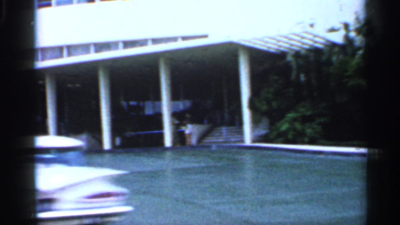 1958 Florida drive-by view of a hotel entrance with white columns, driveway, lawn, and a blurred car crossing the frame.