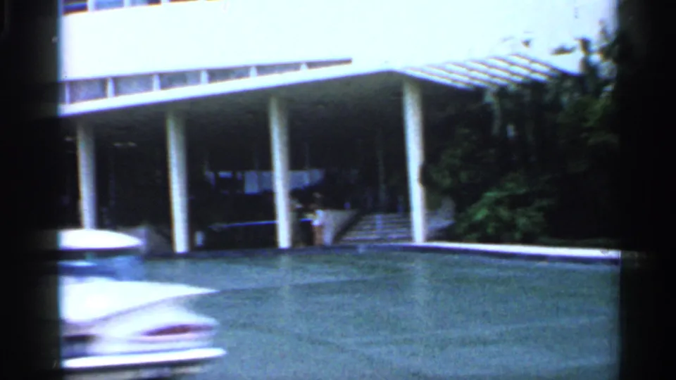 1958 Florida drive-by view of a hotel entrance with white columns, driveway, lawn, and a blurred car crossing the frame.
