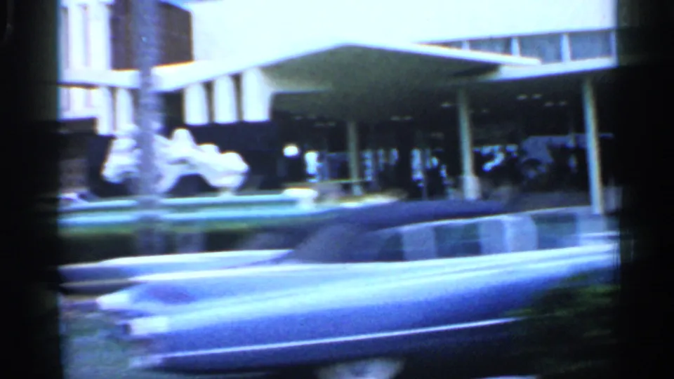 1958 Florida hotel frontage with a covered arrival area, distant people, landscaping, and a classic car blurred in the foreground.