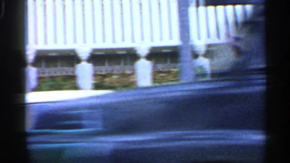 1958 Florida home-movie frame of a hotel exterior with vertical facade panels, lower windows, planting beds, and a passing car.