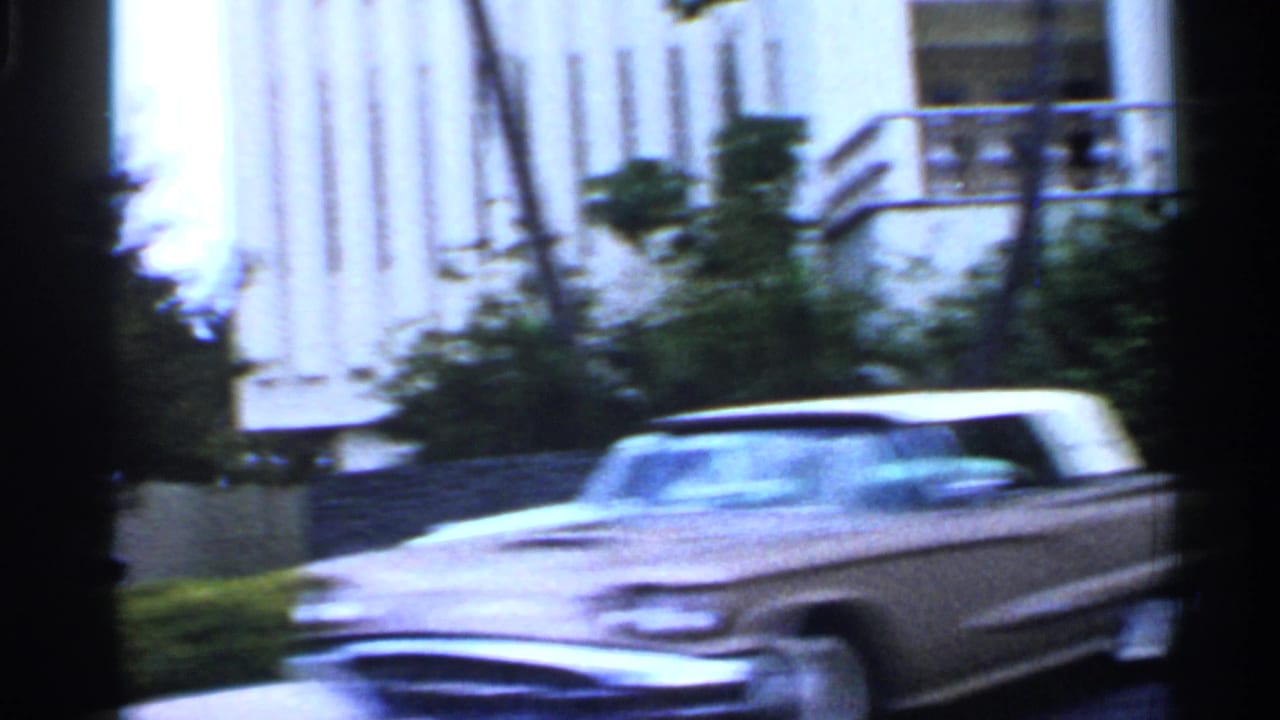 Moving car view past a modern building and a blurred late-1950s car.