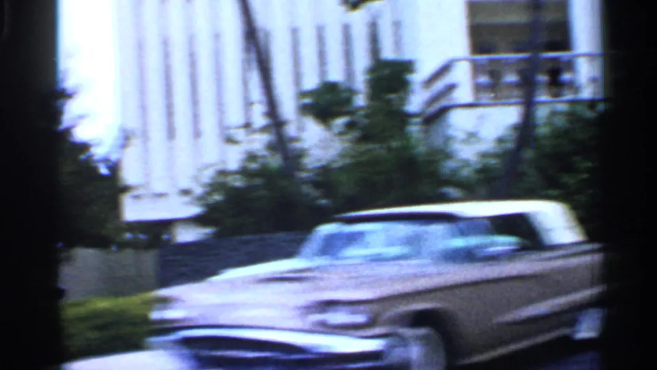 1958 Florida 8mm frame showing a blurred classic car passing a modern building with vertical facade lines and greenery.