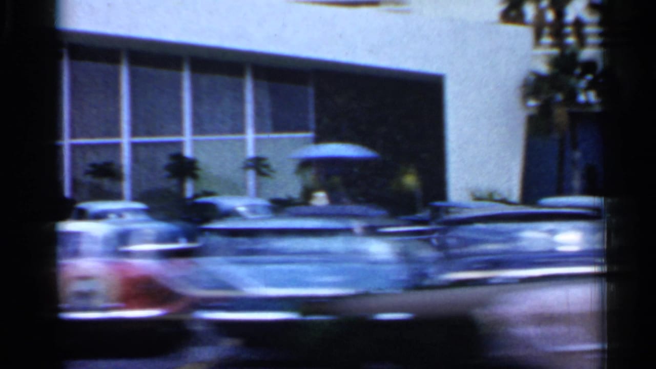 1958 Florida street frame with parked and passing classic cars near a building with large rectangular windows and palms.