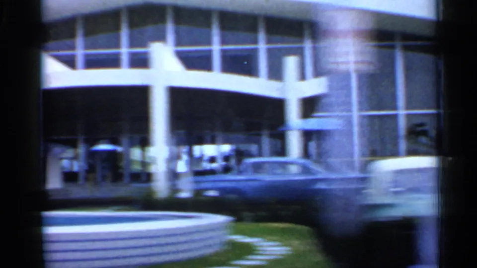 1958 Florida 8mm view of a modern arched entrance, small visible pedestrians, a parked car and a low circular edge in the foreground.