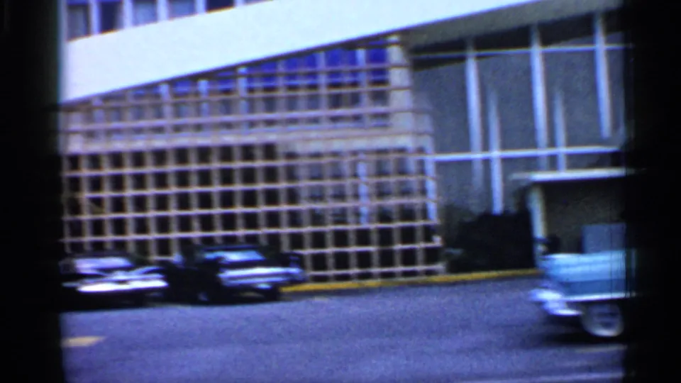 1958 Florida frame showing a gridded modern facade, parked classic cars and a blurred passing car at the edge of the street.