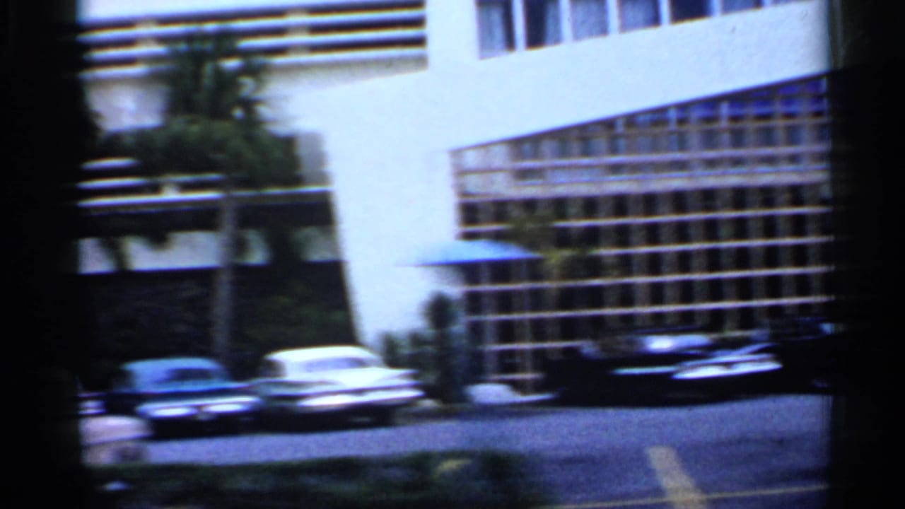 Parked classic cars outside a low-rise mid-century building in bright daylight.
