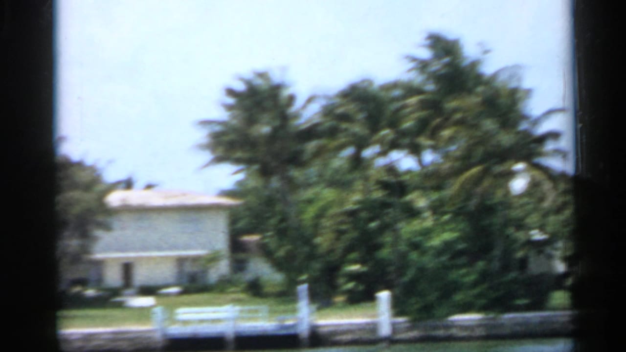 1958 Florida waterfront view with a light-colored house, palm trees, lawn, seawall, and white dock railing.