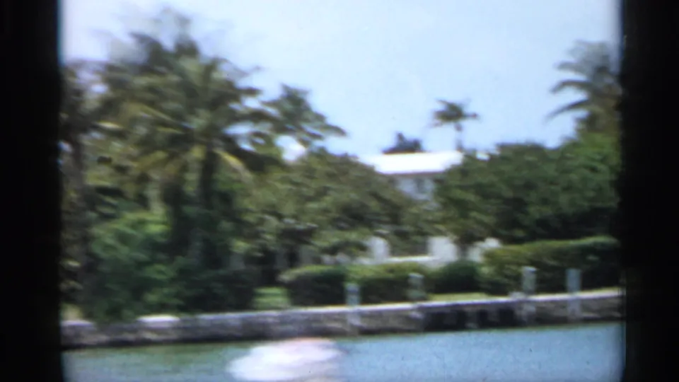 1958 Florida 8mm frame looking across water toward palms, clipped hedges, a seawall, and a pale waterfront home.