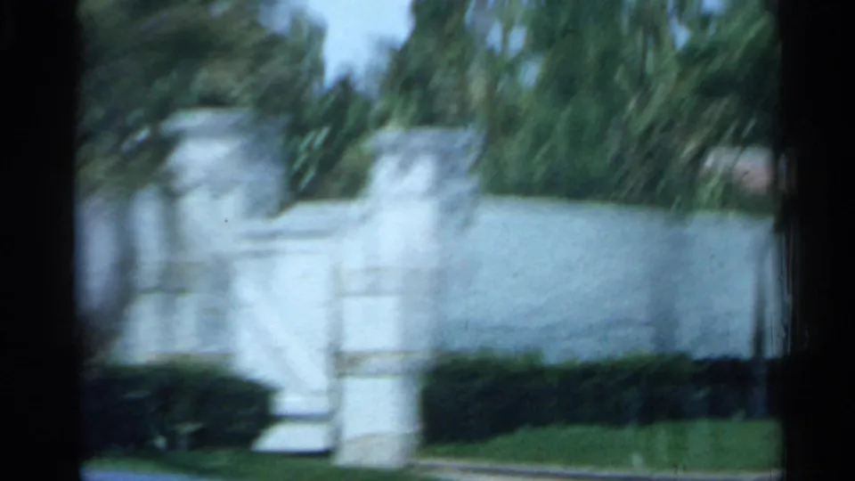 A white Florida building appears beyond hedges and palm branches in a soft, moving 1958 home-movie frame.