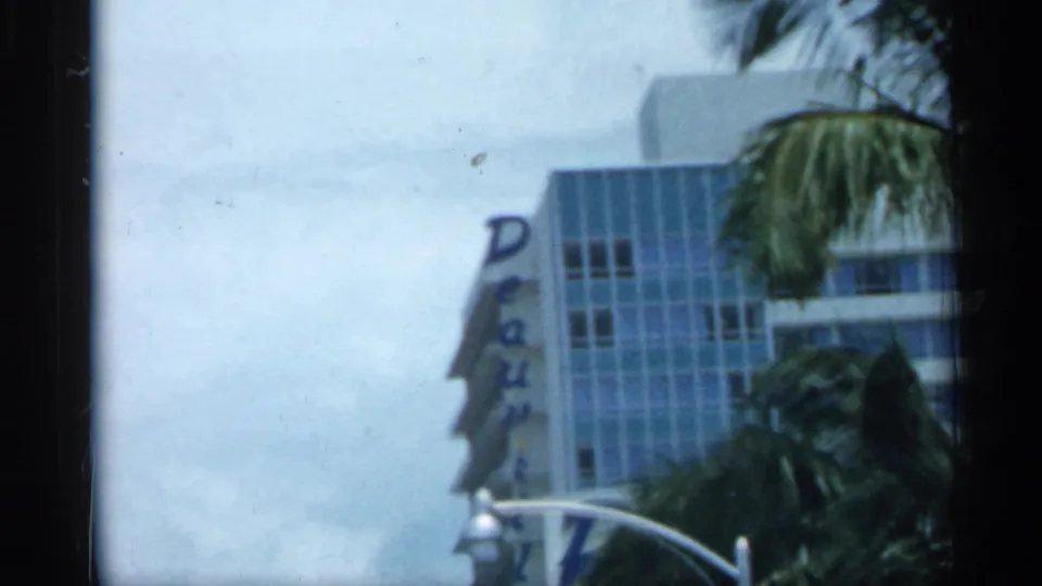 A mid-century Florida building with a visible Deauville sign stands behind palms and cars in a blurred 8mm frame.