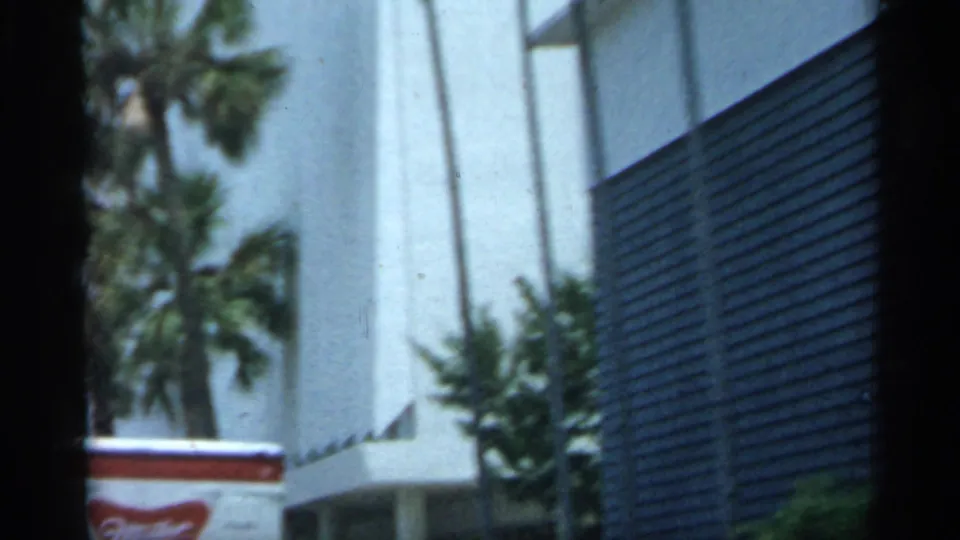 1958 Florida 8mm frame showing palms, a white building, dark shutters, and a red-roofed cart at street level.