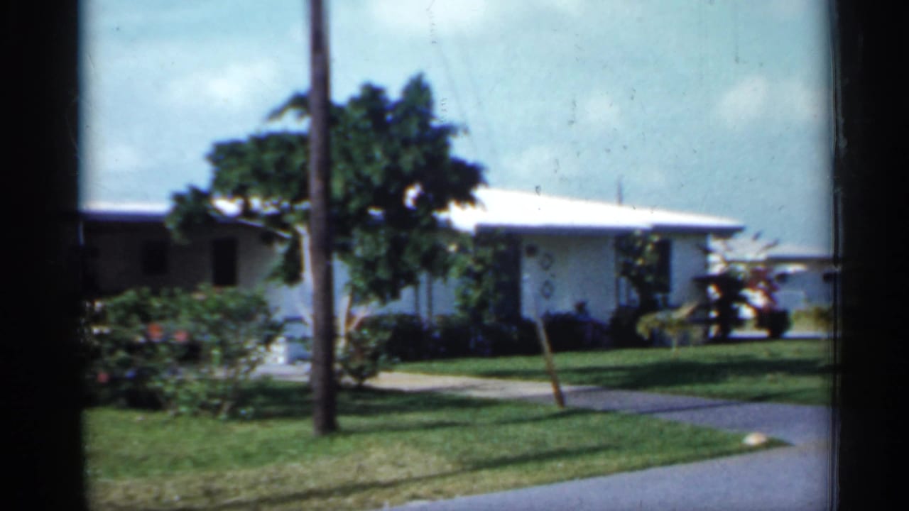 1958 Florida 8mm frame showing a white single-story suburban house with lawn, shrubs, palms, and a roadside view.