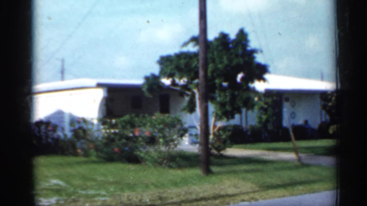 1958 Florida home-movie frame with a white suburban house, broad lawn, palm foliage, shrubs, and a utility pole near the road.