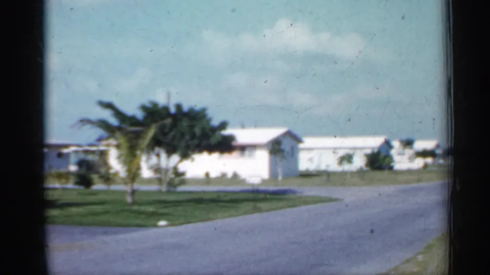 1958 Florida home-movie frame with a curving suburban road, low white houses, open lawns, palm trees, and bright sky.