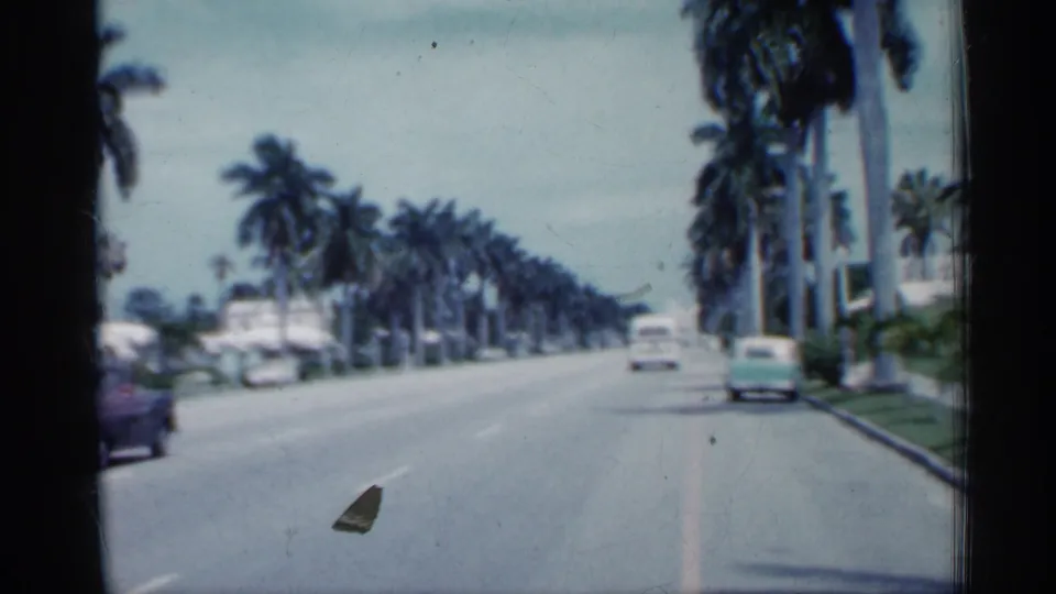 1958 Florida home-movie frame showing a moving view of a palm-lined boulevard with several vintage cars in traffic.