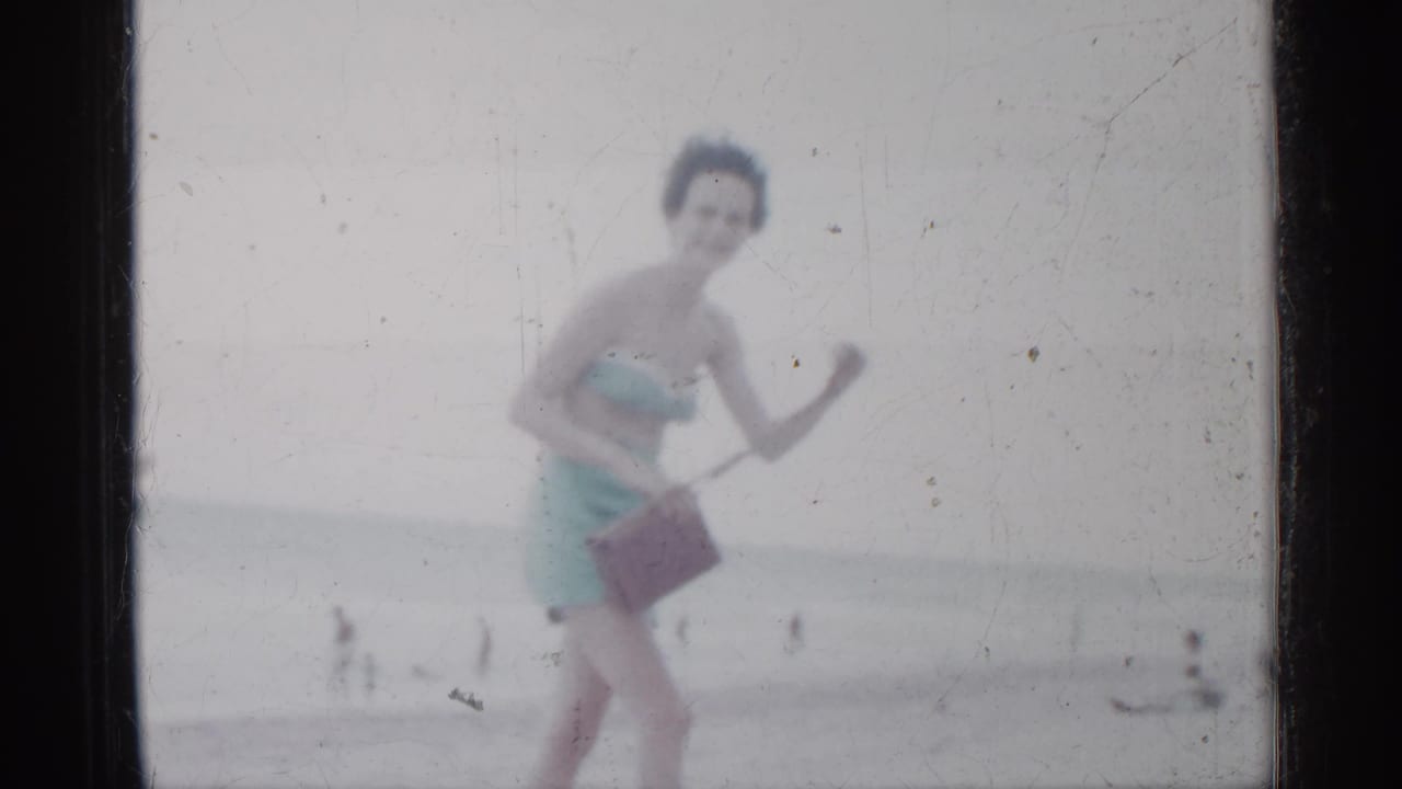 Woman in a light blue swimsuit walks on a beach carrying a brown handbag.