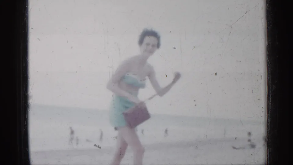 In 1958 Florida home-movie footage, a woman in a light blue swimsuit smiles while walking on the beach with a brown handbag.