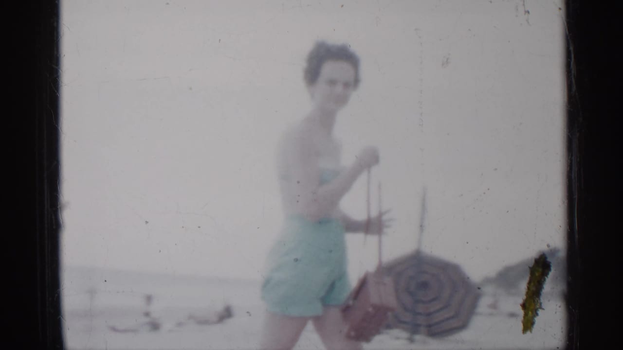 The woman in a light blue swimsuit pauses near a striped umbrella on a Florida beach, with distant swimmers and horizon behind her.