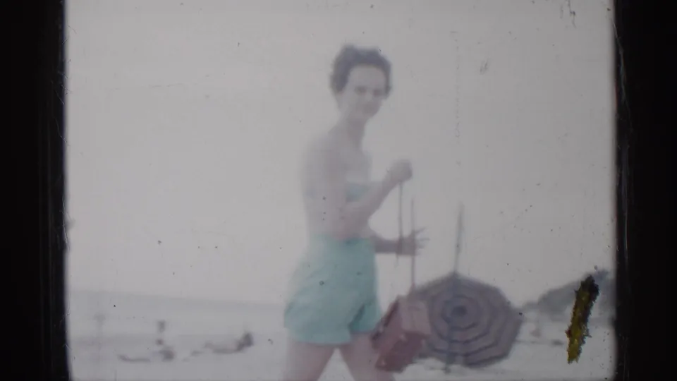 The woman in a light blue swimsuit pauses near a striped umbrella on a Florida beach, with distant swimmers and horizon behind her.