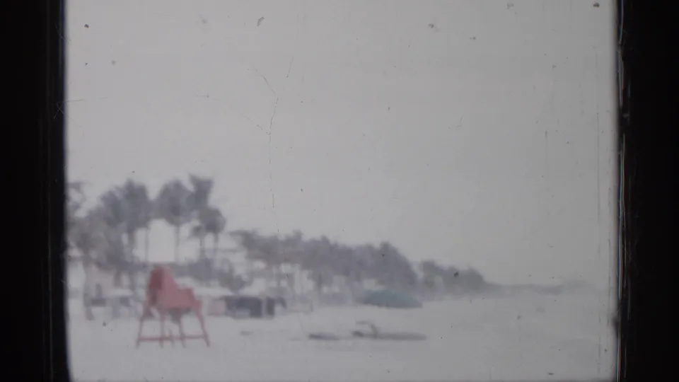 1958 Florida 8mm frame showing windblown palms, beach sand, water, and a red lifeguard chair in stormy weather.