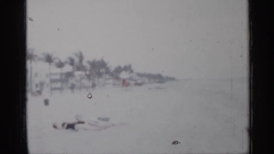 1958 Florida 8mm frame showing a person low on the beach while palms, buildings, and shoreline recede into storm haze.