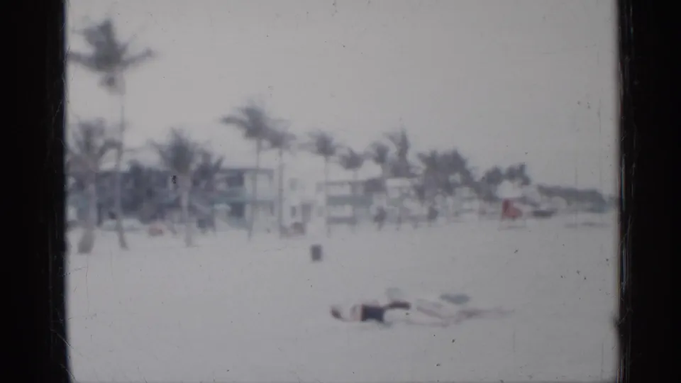 1958 Florida storm beach frame with a person on the sand, windblown palms, low buildings, and a red lifeguard chair in view.