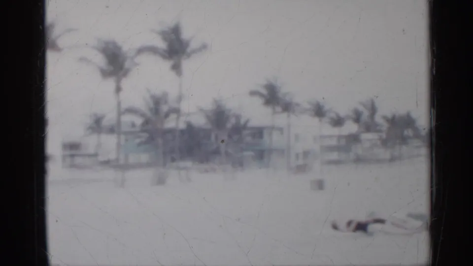 1958 Florida beach view with palms, pale sand, low-rise buildings, and one distant beachgoer lying near the lower center.