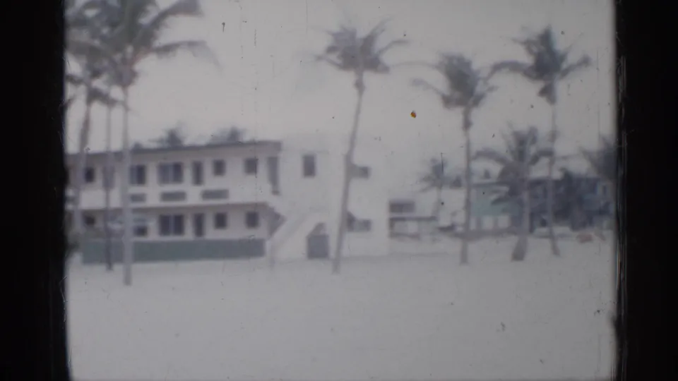 1958 Florida shoreline building framed by palm trees, pale sand, and a visible exterior stairway in soft 8mm focus.