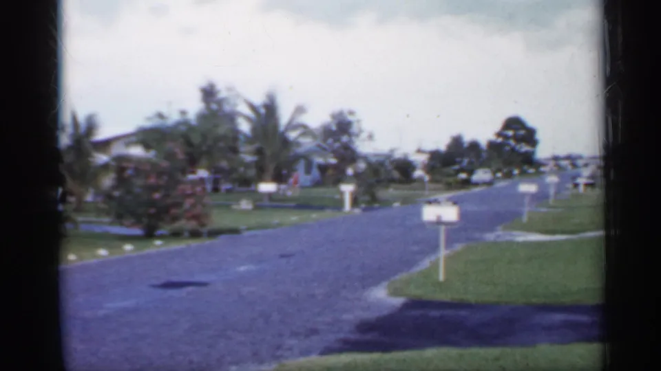 1958 Florida 8mm frame showing a quiet suburban street with green lawns, curbside mailboxes, palm trees, and low homes.