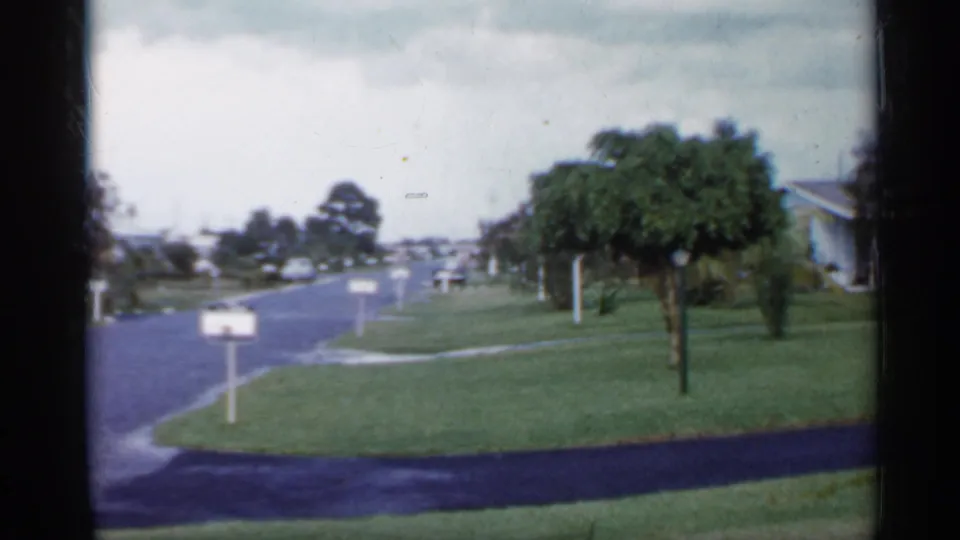 1958 Florida 8mm view of a suburban street edge with a mailbox, trimmed grass, a small tree, houses, and distant people.