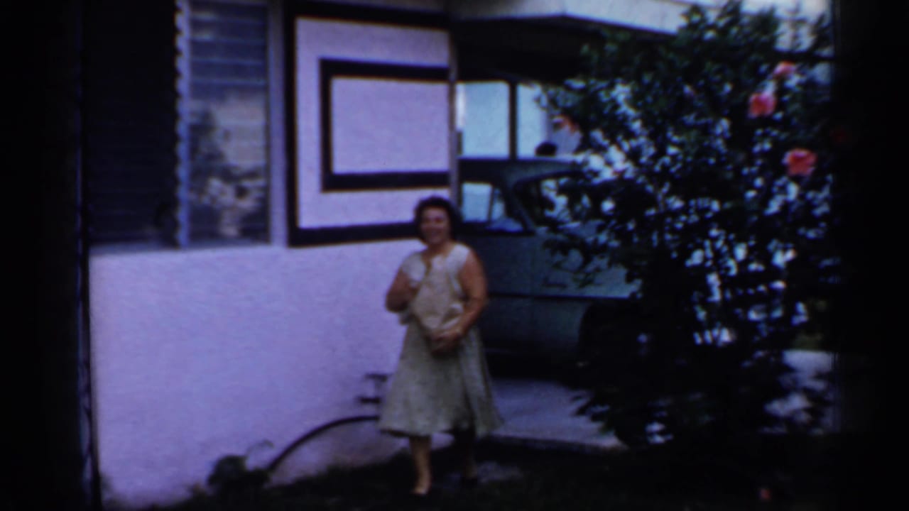A woman stands outside a Florida mobile home in 1958, holding a paper bag near the entrance with a parked car partly visible.