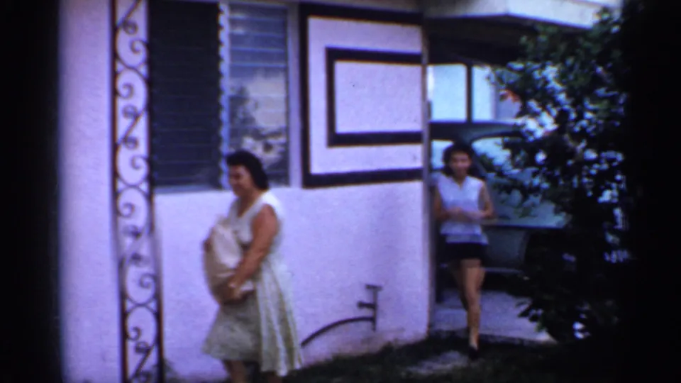 Outside the mobile home, a woman holds a paper bag near decorative porch ironwork while another person stands by a parked car.