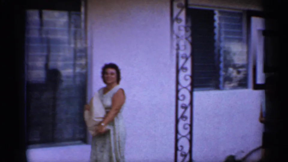 The woman faces the camera beside the mobile home wall, holding a paper bag near a window and decorative porch support.