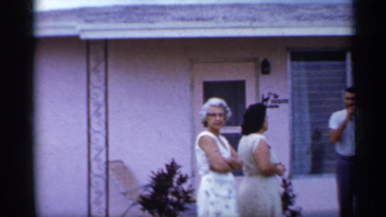 1958 Florida home-movie frame showing two women and a man outside a pink stucco house during a casual neighborhood conversation.